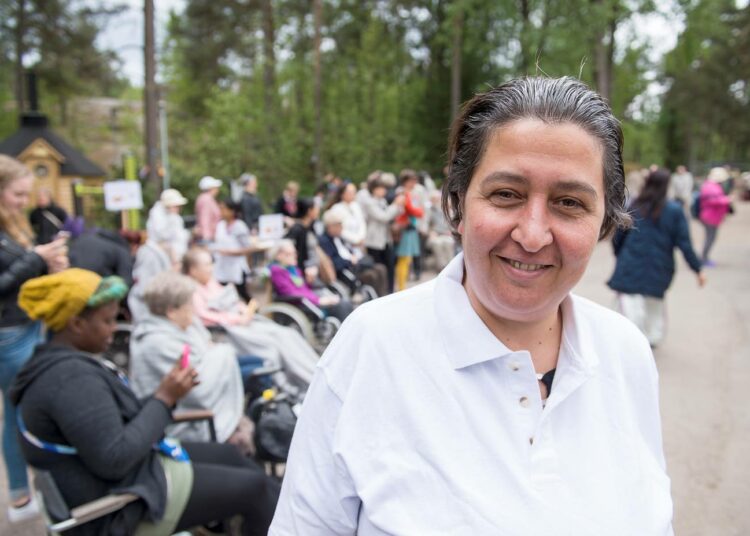 Mania Alkhatib on Suomi–Syyria-ystävyysseuran puheenjohtaja ja aktiivinen Riistavuoren vanhusten kanssa tehtävässä vapaaehtoistyössä. Taustalla yleisöä seuraamassa suomalais-syyrialaisen kuoron esiintymistä Riistavuoren palvelukeskuksen monikulttuurisena iltapäivänä.