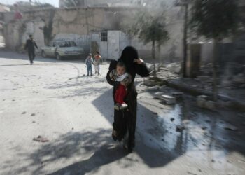 Äiti juoksee lapsi sylissään suojaan ilmahyökkäykseltä Kafr Batnan kaupungissa.