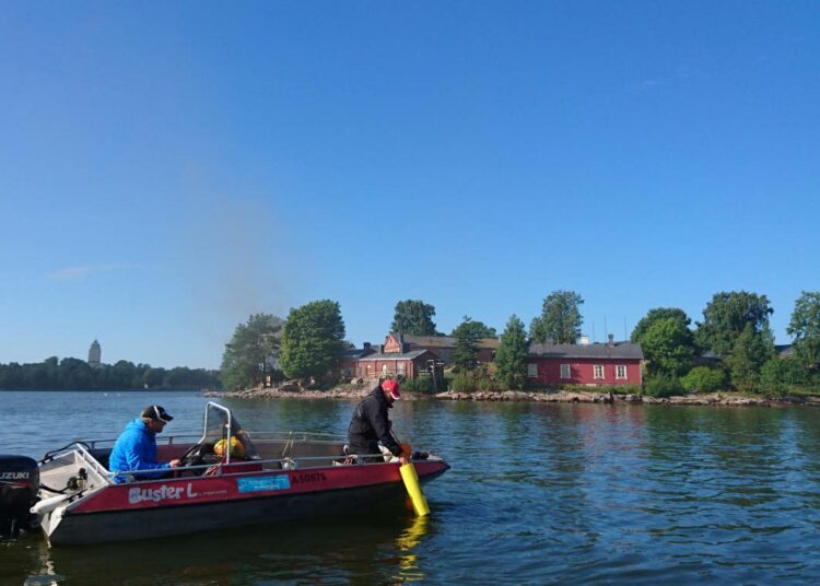 Miska Kopponen Helsingin kaupungin kulttuurin ja vapaa-ajan toimialalta ja Heikki Peltonen Suomen ympäristökeskuksesta nostavat mittalaitteita Kruunuvuorenselällä. Taustalla Lonna ja Suomenlinna.