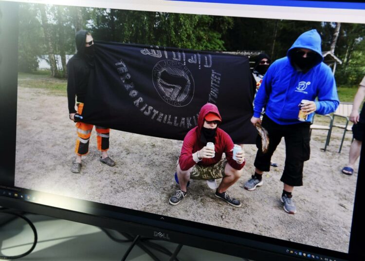 Perussuomalaisten kesäpäivillä Keuruulla 2018 otetussa kuvassa neljä kasvonsa peittänyttä miestä poseeraa juutalaisvastaisen lipun kanssa. Lipun keskellä lukee anti judische aktion.